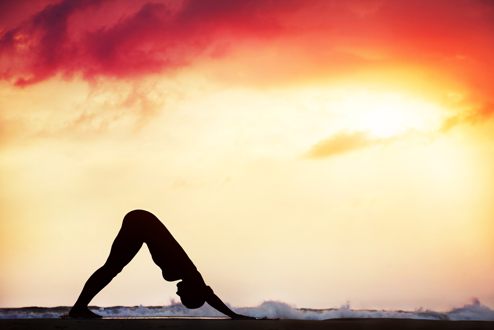 Yoga on vacation.  Yoga While Traveling.
Step of surya namaskar, downward facing dog pose by beautiful woman on the beach near the ocean at dramatic sunset background