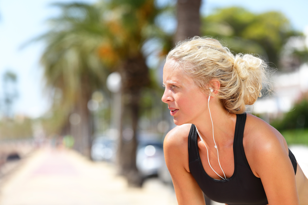 Summer Vacation Safety Tips.  Tired running woman taking a break during run. Beautiful young blond athletic female adult resting catching her breath while jogging and listening to music with earphones and smartphone app.