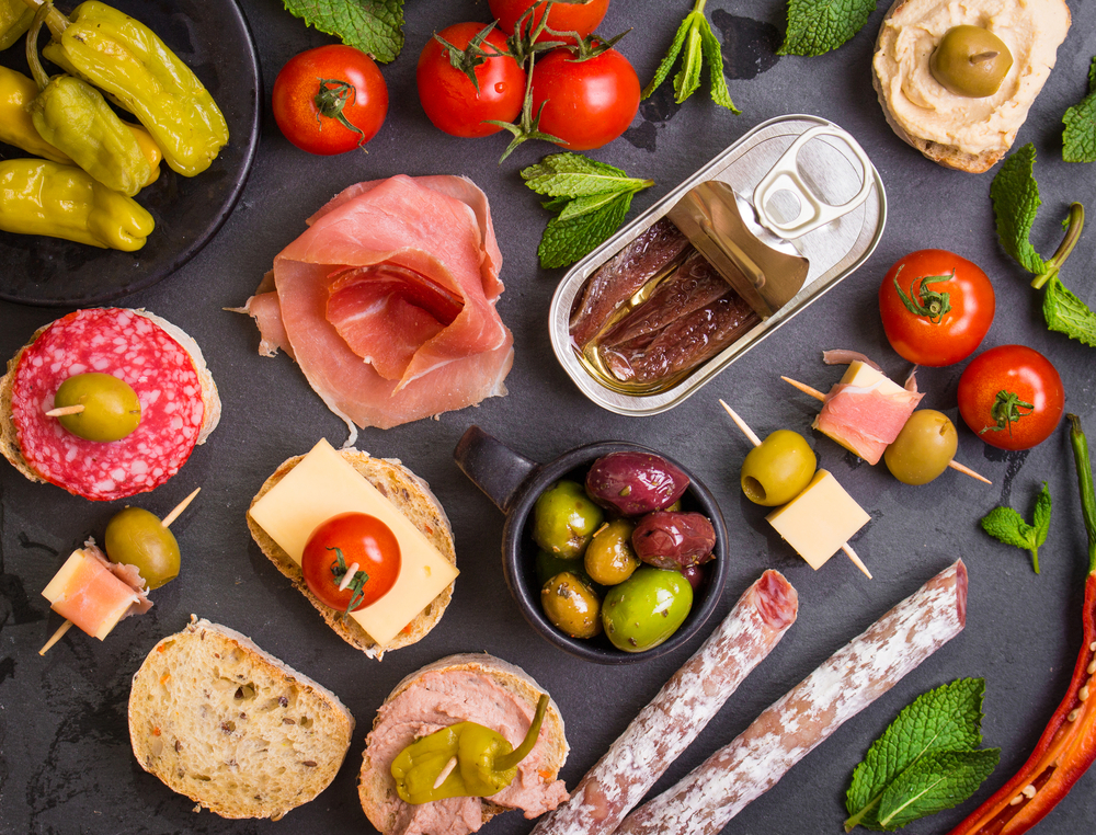 Summer stay cation ideas.  Create a charcuterie board with a 
Mix of different snacks and appetizers. Spanish tapas on a black stone plate. Tapas bar. Deli, sandwiches, olives, sausage, anchovies, cheese, jamon, marinated pepper, tomatoes, canapes. Top view