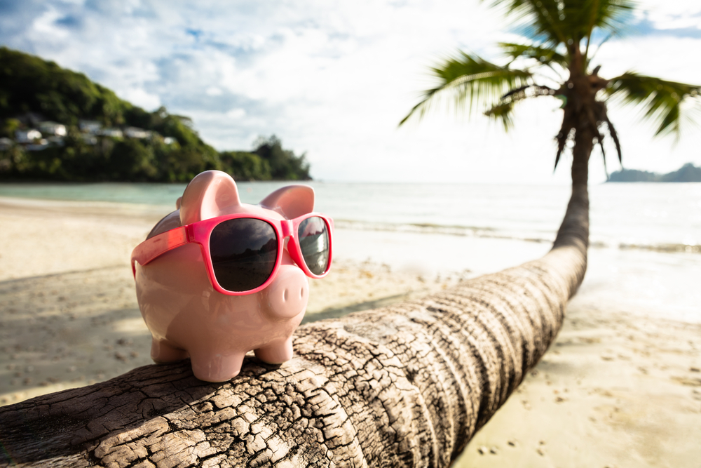 Close-up Of Pink Piggybank With Sunglasses On Tree Trunk At Beach.
How to Build a Travel Fund.
