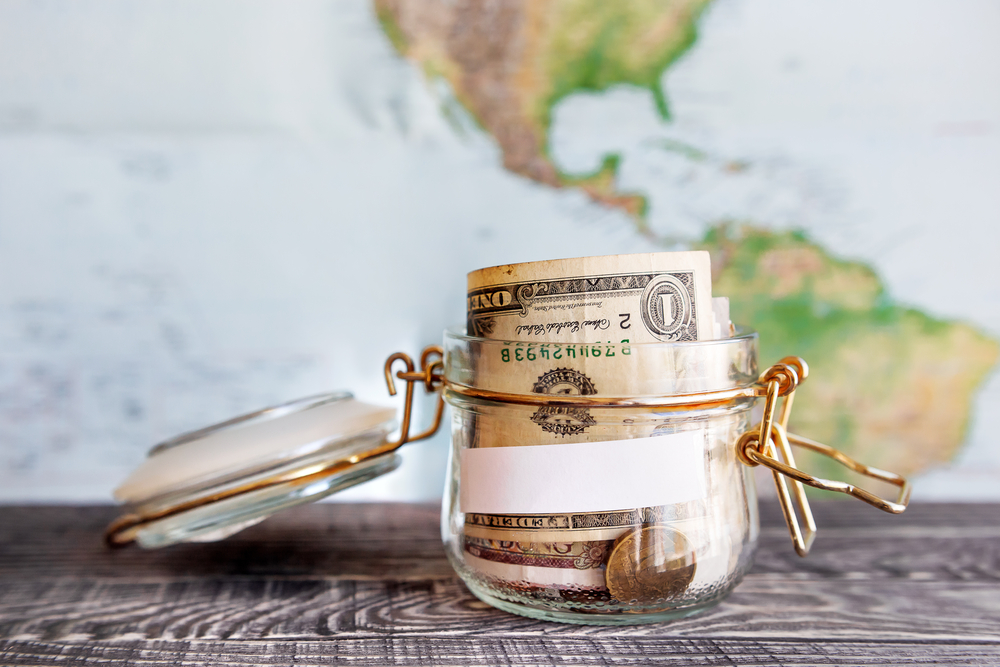 How to Build a Travel Fund.  Jar of cash with a map backdrop.