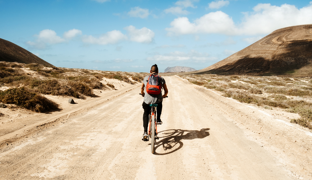 Sustainable Travel Kit.
A young man seen from behind riding a bike in a dirty road in La Graciosa, Canary Islands, Spain
