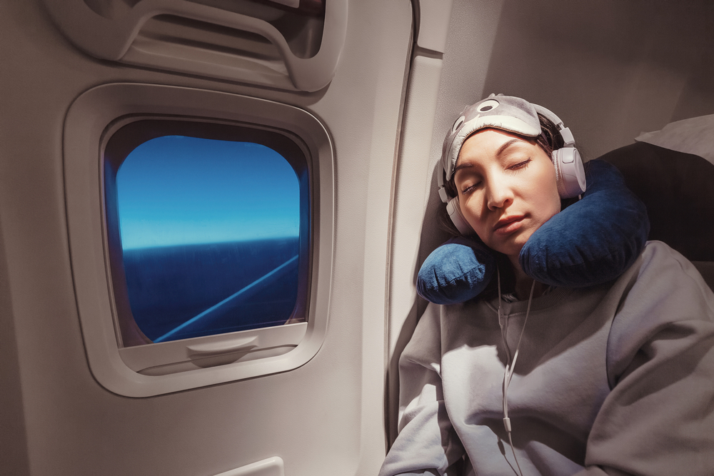 How to Get Over Jet Lag.
Girl sleeping in her seat on the plane near the window in a mask and with a pillow to sleep. The concept of travel with comfort and jetlag