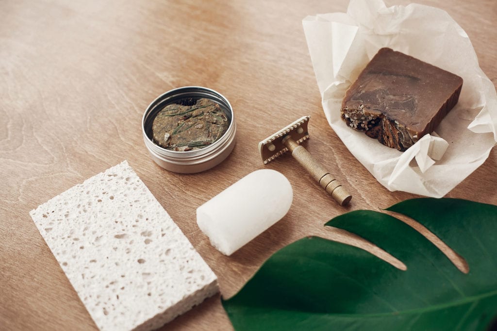 Eco-Friendly Travel Products.
Zero waste, plastic free beauty essentials. Natural soap, solid shampoo in metal tin, reusable razor, crystal eco deodorant, sponge on wooden background with green monstera leaf.