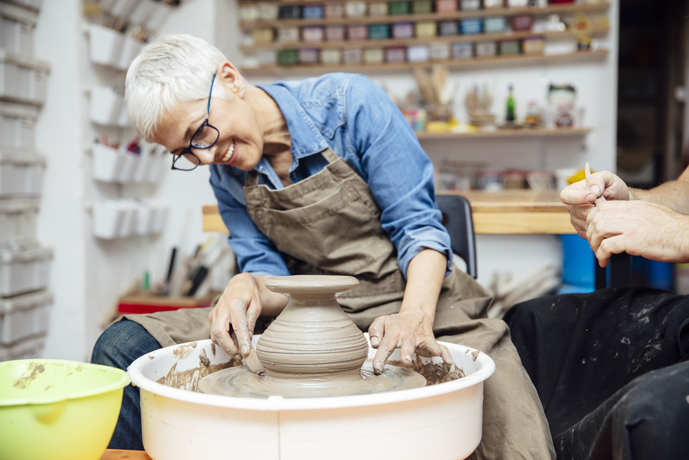 60 Things to do Before 60.
Woman spinning clay on a wheel with a help of a teacher at pottery class